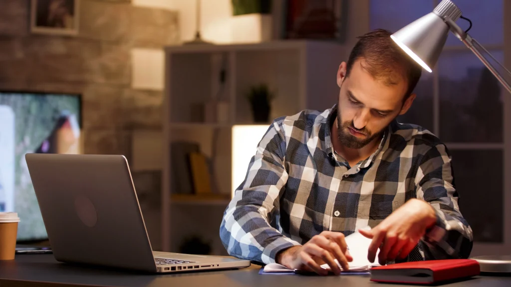 Man working late at desk with laptop and notebook under lamp