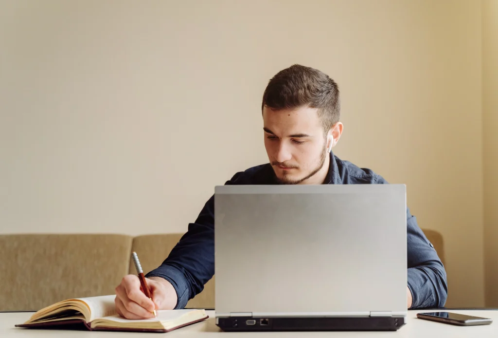Man writing notes while using laptop at desk
