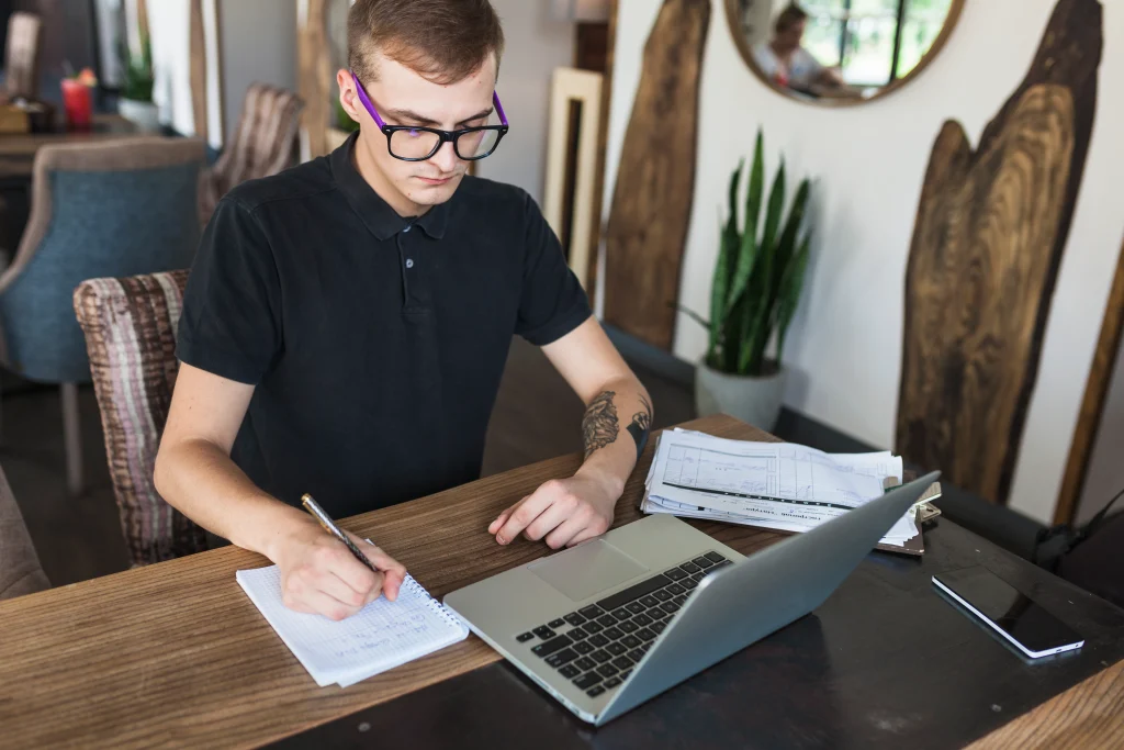 Young male student studying online, taking notes while using laptop