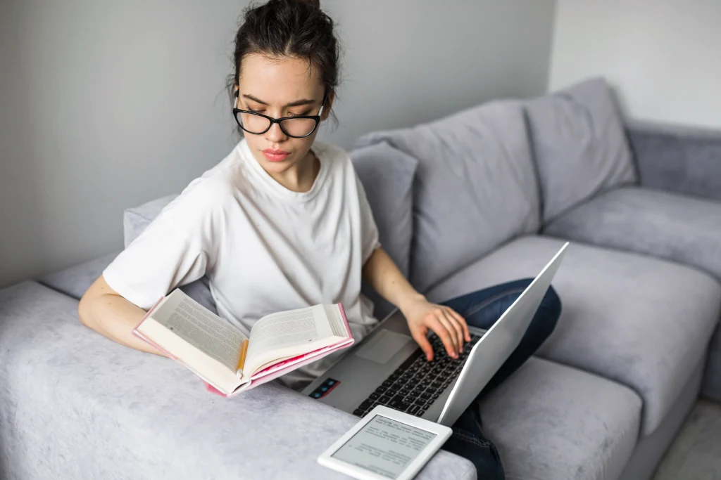woman reading book and using laptop on sofa