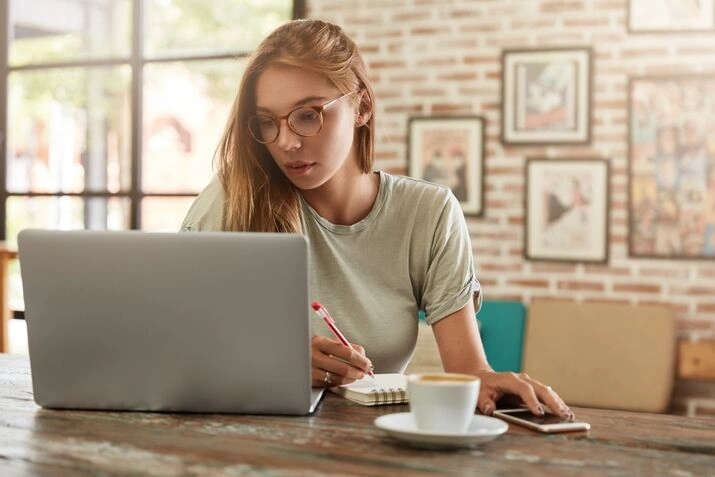 Woman writing notes while working on laptop at home office