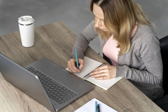 Woman making notes beside laptop on wooden workspace