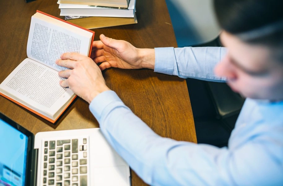 person reading book while working on laptop desk